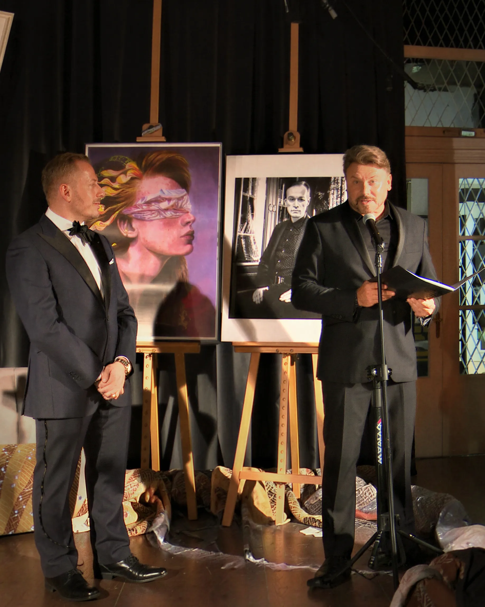 Two hosts in evening wear speaking on stage beside framed artworks